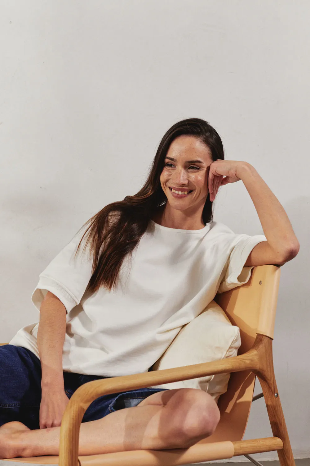 Woman sits smiling on a chair and supports her arm. The background is calm and neutral.