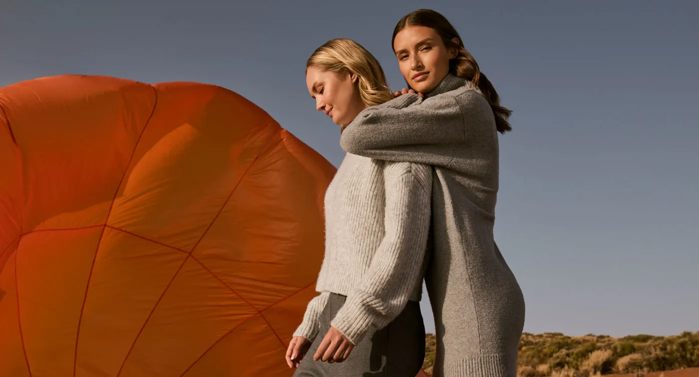 Zwei Frauen in warmen Strickpullovern stehen vor einem orangefarbenen Heißluftballon in einer herbstlichen Landschaft.
