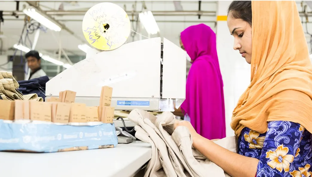 Eine Mitarbeiterin sitzt konzentriert am Tisch und arbeitet an einem beigefarbenen Kleidungsstück. Im Hintergrund sind weitere Mitarbeiterinnen und die Fabrik zu sehen.