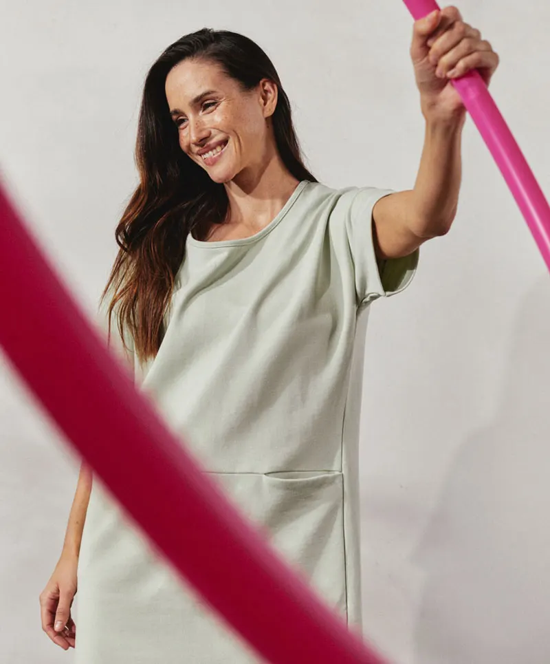 A woman in a light green dress holds a pink hula hoop. She looks to the right, smiling. The background is neutral.