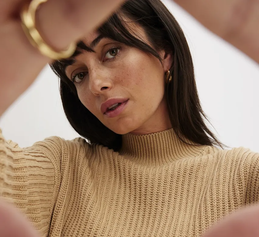 Eine Frau mit dunklem Haar in einem beigen Pullover blickt nachdenklich zur Seite.