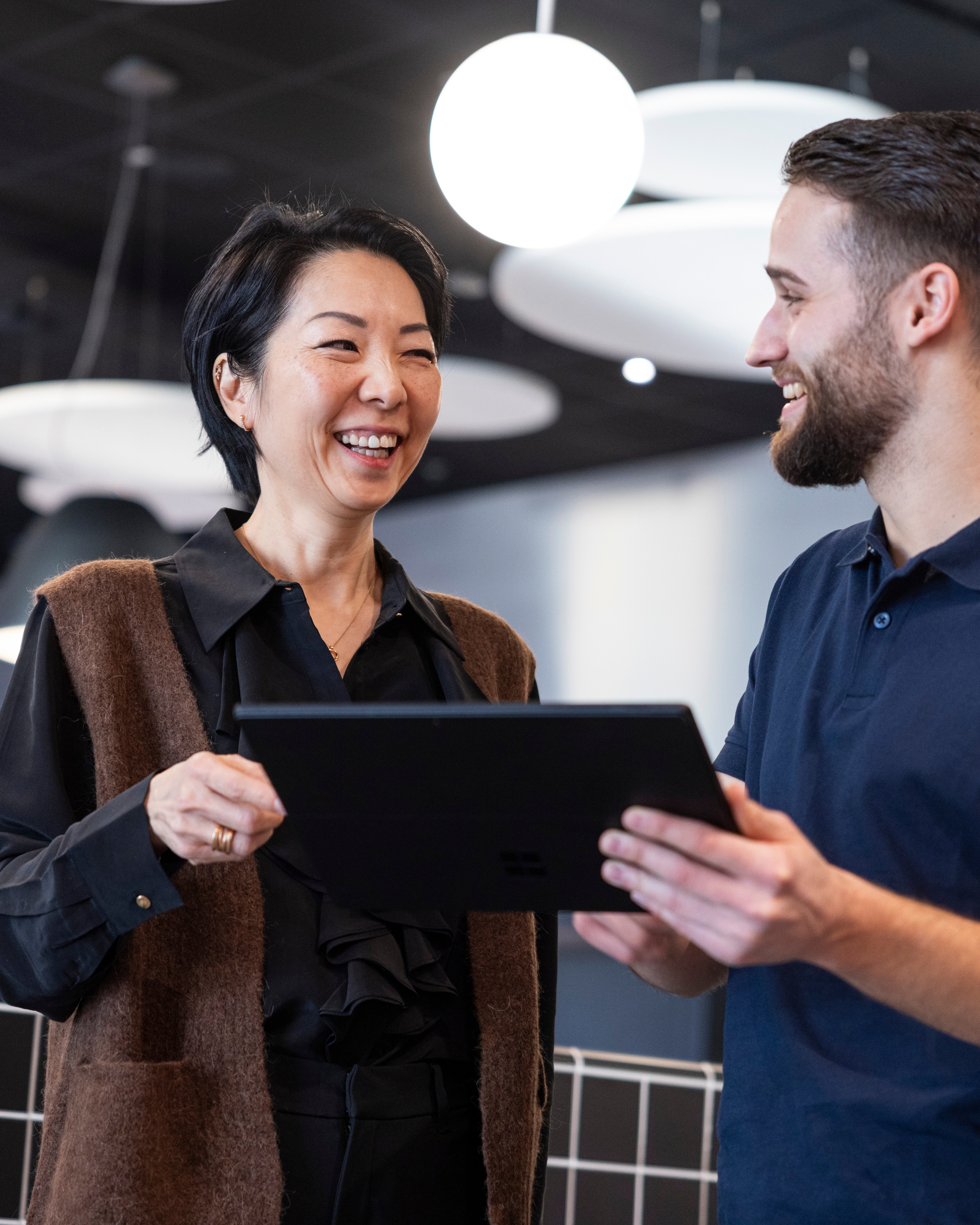 Ein lächelndes Paar in warmen Pullovern steht vor einer Wand mit Fotos.