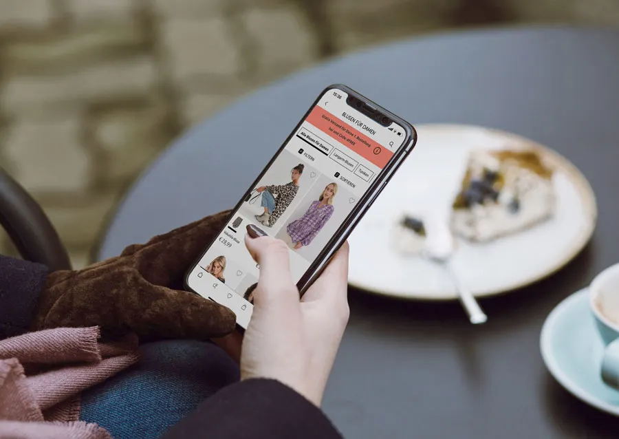 Smartphone zeigt Online-Shopping-App. Daneben Kaffeetasse und Teller mit Dessert auf Café-Tisch.