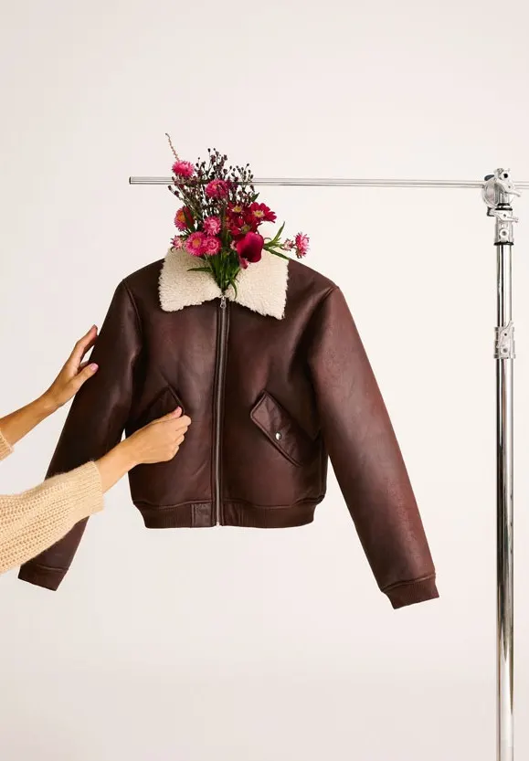 A brown jacket with a fake-fur collar hangs from a coat rack. A bouquet of flowers hangs from the collar. Hands touch the jacket.