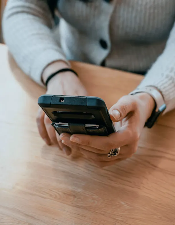 Hände halten Smartphone auf Holztisch. Grauer Pullover und silberner Ring sind sichtbar.