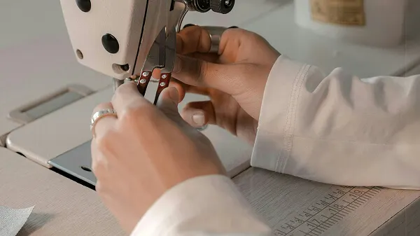 Nahaufnahme von Händen, die an einer weißen Nähmaschine arbeiten und eine kleine Fadenschere im Bereich der Nadel halten.
