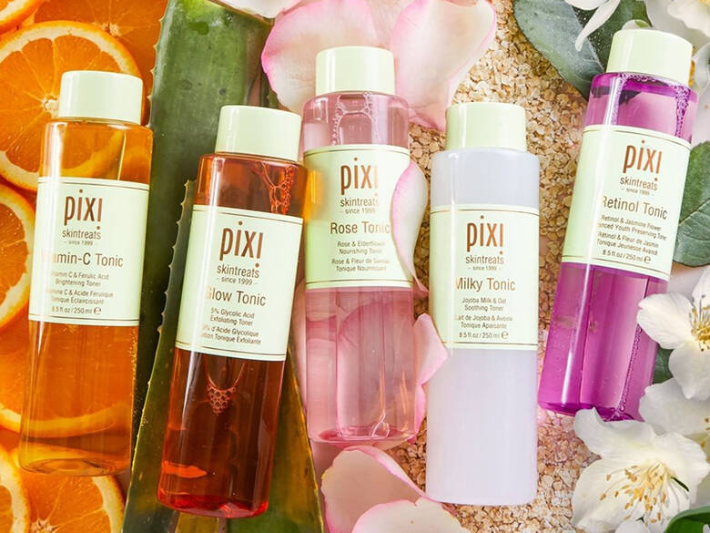 Her ses fem Pixi Beauty produkter i lyserøde og orange nuancer. De ligger på forskellige planter.