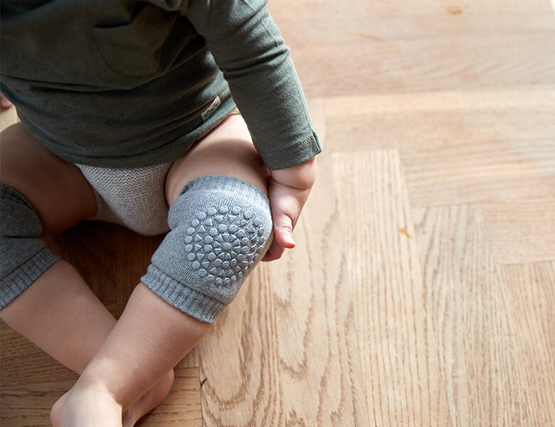 Her ses en baby sidde på et trægulv. Babyen har grå knæbeskyttere på.