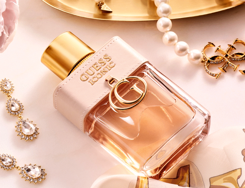 Her ses et billede af en parfumeflakon med teksten “GUESS ICONIC” placeret på en lys overflade. Flakonen er i klare og rosa nuancer med guldfarvet låg og detaljer. Omkring flakonen ses smykker som perler og øreringe.