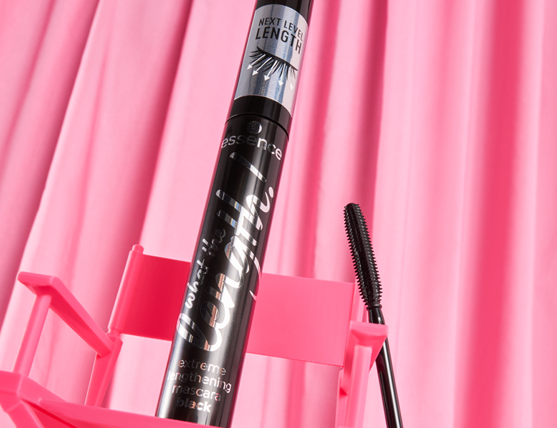 Her ses et billede af en mascara fra essence placeret mod en pink baggrund med folder. På produktet står teksten “essence what the fake! extreme lengthening mascara black” og “NEXT LEVEL LENGTH”. En mascarabørste er vist ved siden af.
