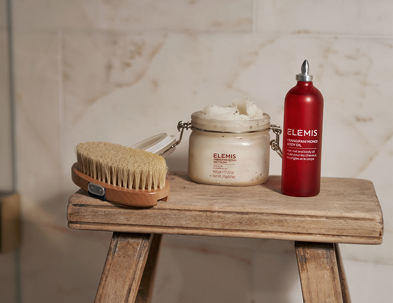 Her ses ELEMIS kropsplejeprodukter – body olie, scrub og børste placeret på en træskammel.