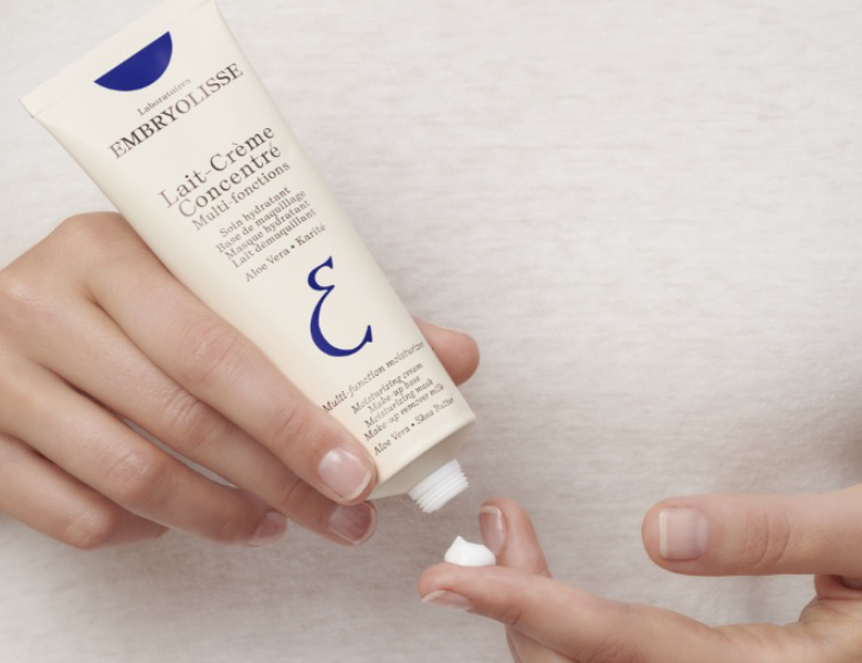 Her ses en hånd, der holder en tube Embryolisse Lait-Crème Concentré, mens creme påføres en finger, mod en lys baggrund.