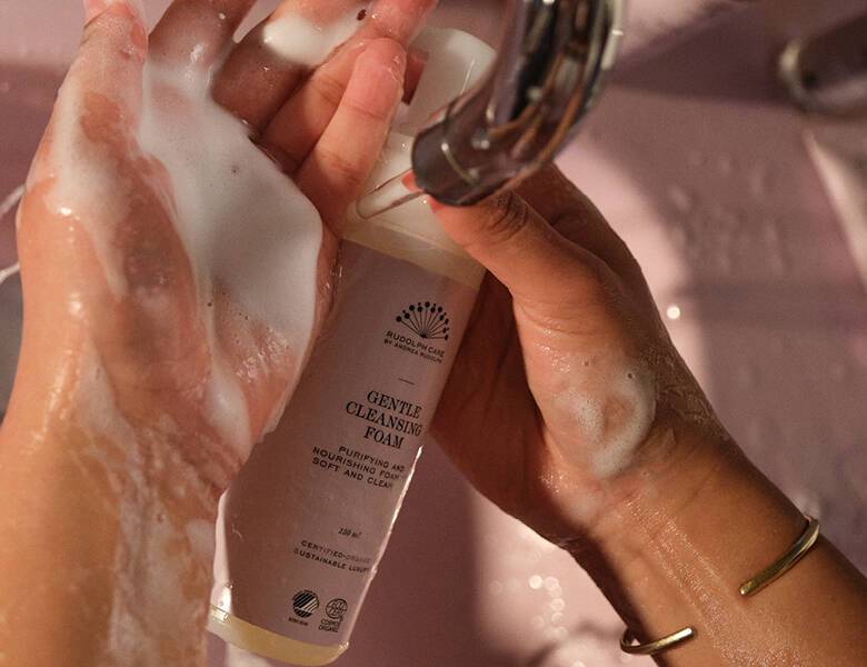 Her ses nogle hænder under en vandhane. Den ene hånd holder produktet og den anden hånd har selve produktet på. Håndvasken er lyserød. 