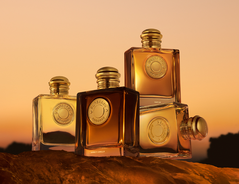 Her ses et billede med fire parfumeflakoner fra Burberry Goddess placeret mod en baggrund med varme solnedgangsfarver. Flakonerne vises i forskellige størrelser og varianter, herunder en mørkere udgave med teksten “Amber Vanilla” og en større flakon med teksten “Parfum”.