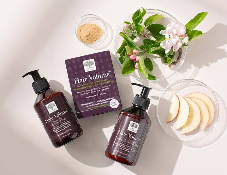 Her ses en lys baggrund med tre lilla New Nordic produkter blandt nogle blomster og skiveskåret æbler. 