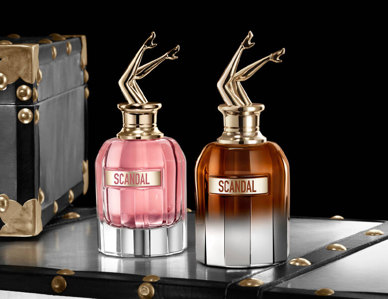 Her ses et billede med to parfumeflakoner med teksten “SCANDAL” på fronten. Den ene flakon er lyserød, og den anden er brunlig med en gradient mod klar bund. Begge har låg formet som et par opadvendte ben i guldfarve. Produkterne er placeret på en mørk overflade med dekorative kuffertelementer i baggrunden mod en sort baggrund.