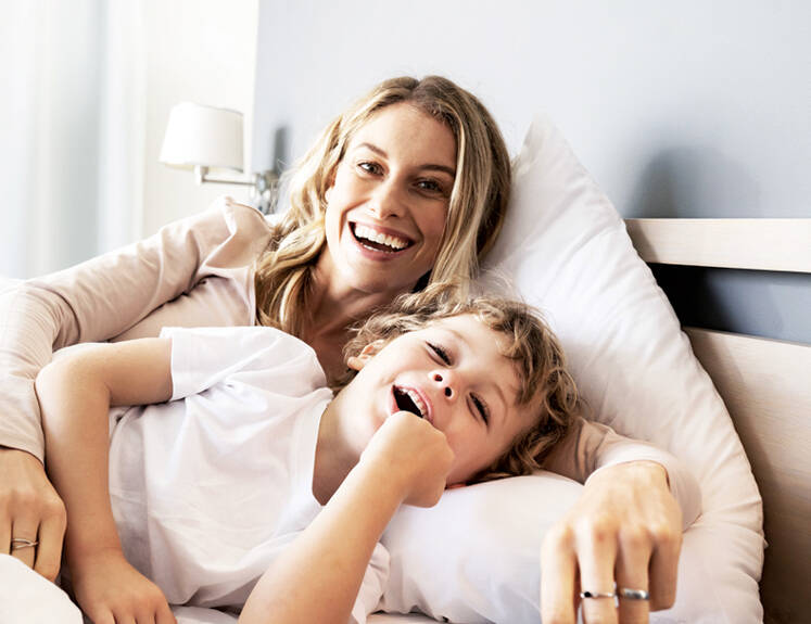 Her ses en glad mor og sin søn. De ligger i en seng. 
