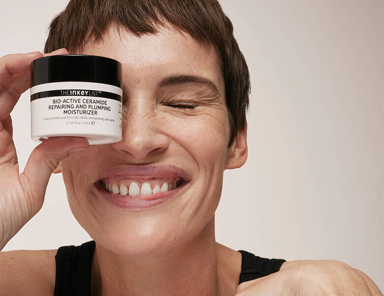 Her ses en grå baggrund med en kvinde, som holder en creme fra The INKEY List foran ansigtet.