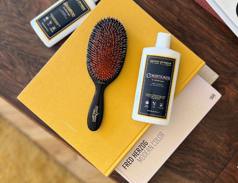 Her ses en sort hårbørste liggende på en gul bog med et hvidt produkt ved siden af. Bordet er af mørkt træ. 