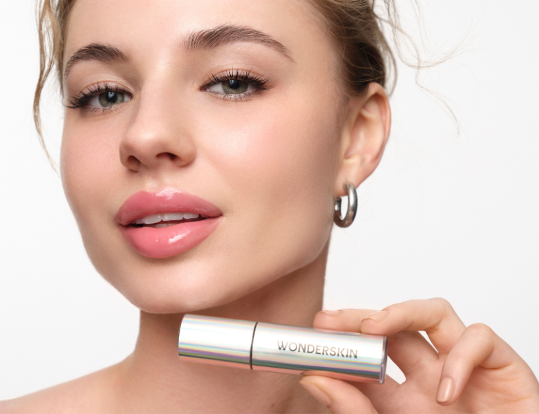 Her ses et nærbillede af en person, der holder en Wonderskin-læbeprodukt op foran ansigtet. Personen har blanke, lyserøde læber og et naturligt makeup-look, mens produktet fremhæves som fokus i billedet.