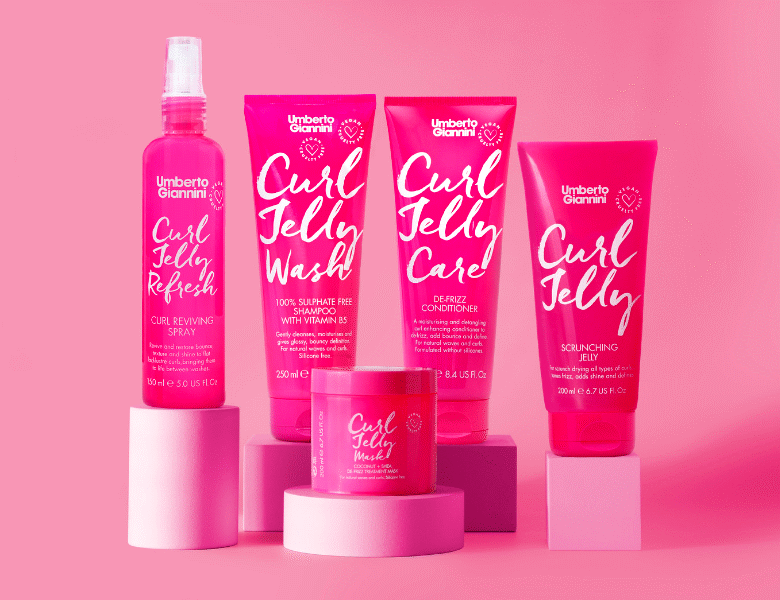 Her ses et billede med flere hårprodukter fra Umberto Giannini placeret på små podier mod en lyserød baggrund. På produkterne står blandt andet “Curl Jelly Refresh”, “Curl Jelly Wash”, “Curl Jelly Care” og “Curl Jelly”. Produkterne er i pink emballage med hvid tekst.