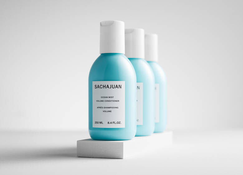 Her ses et billede med flere flasker fra SACHAJUAN placeret på række mod en lys baggrund. På den forreste flaske står teksten “SACHAJUAN Ocean Mist Volume Conditioner”. Flaskerne er i lyseblå farve med hvide låg.
