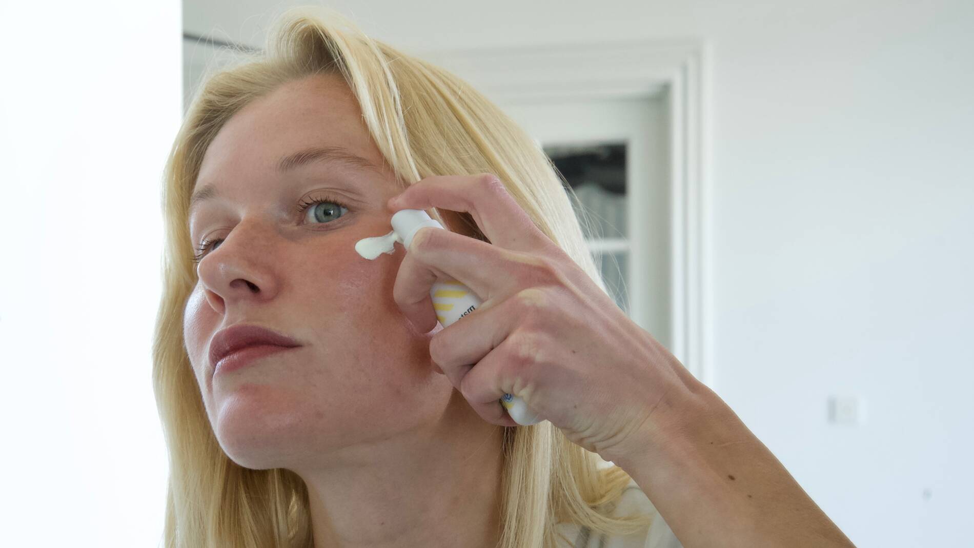 En person med lyst hår påfører ansigtscreme på kinden med fingrene, mens personen holder en flaske med solcreme fra Matas Striber i den anden hånd. Personen står indendørs foran et spejl i et lyst badeværelse og ser fokuseret mod spejlet. Solcremen har hvid emballage med gule striber og flere certificeringsikoner nederst på flasken.