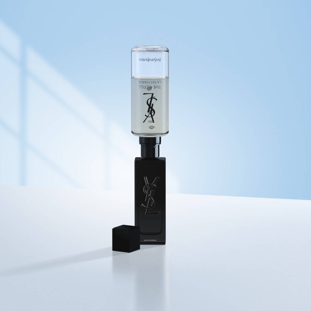 En sort Yves Saint Laurent MYSLF-parfumeflaske står på en lys overflade foran en blød lyseblå baggrund. Oven på flasken er en gennemsigtig refillbeholder monteret for at vise genopfyldning af parfumen. Flaskens sorte låg ligger ved siden af. Billedet har et stilrent og eksklusivt udtryk med fokus på luksusparfume og refill-løsninger.
