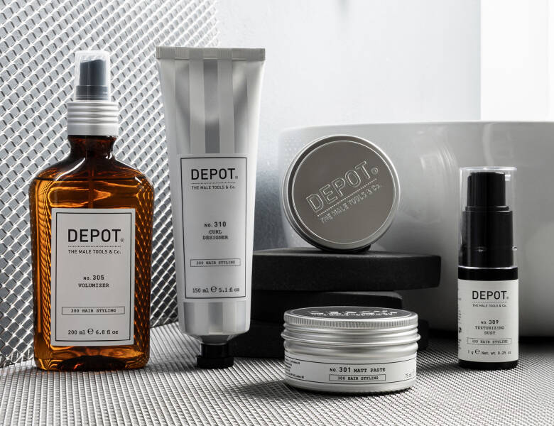 Her ses et billede med flere produkter fra DEPOT placeret på en overflade ved en vask. På produkterne står blandt andet “DEPOT”, “No. 305 Hair Styling”, “No. 310 Curl Designer”, “No. 301 Matt Paste” og “No. 309 Texturizing Dust”. Produkterne er i brune og sølvfarvede emballager med hvide etiketter.