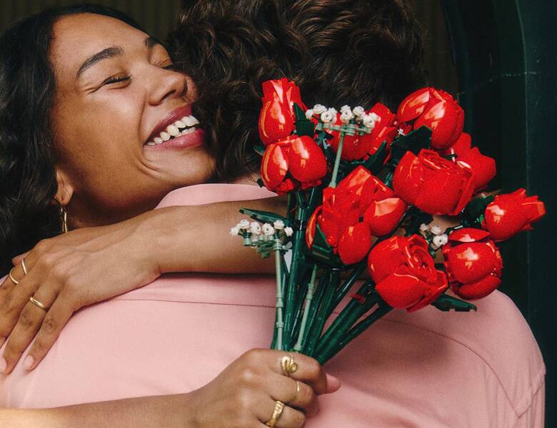 Her ses en kvinde som krammer en mand i en lyserød skjorte. Hun er meget glad og holder en buket af røde Lego-blomster. 