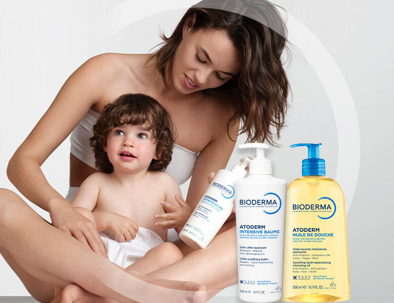 Her ses en mor, som sidder med sit barn i skødet. Moren holder et BIODERMA produkt i hånden og foran dem vises to BIODERMA produkter. 