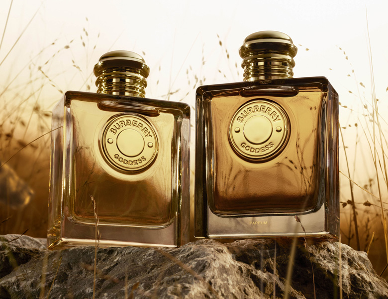 Her ses et billede med to parfumeflakoner fra Burberry placeret side om side på en sten i et landskab med græs. På flakonerne står teksten “BURBERRY GODDESS”. Flakonerne er i gyldne nuancer med runde detaljer på fronten.