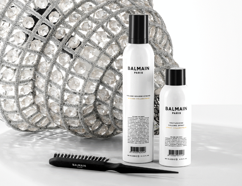 Her ses en hvid baggrund med en lysekrone. Foran ses en styling-hårbørste samt to hvide og sorte produkter. 