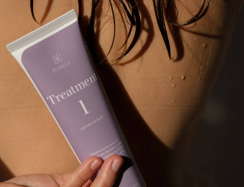 Her ses et billede med en person med vådt hår, hvor et produkt holdes op foran kroppen. På produktet står teksten “PURELY Treatment 1” samt “LEAVE-IN HAIR”. Der ses vanddråber på huden, og baggrunden er mørk.
