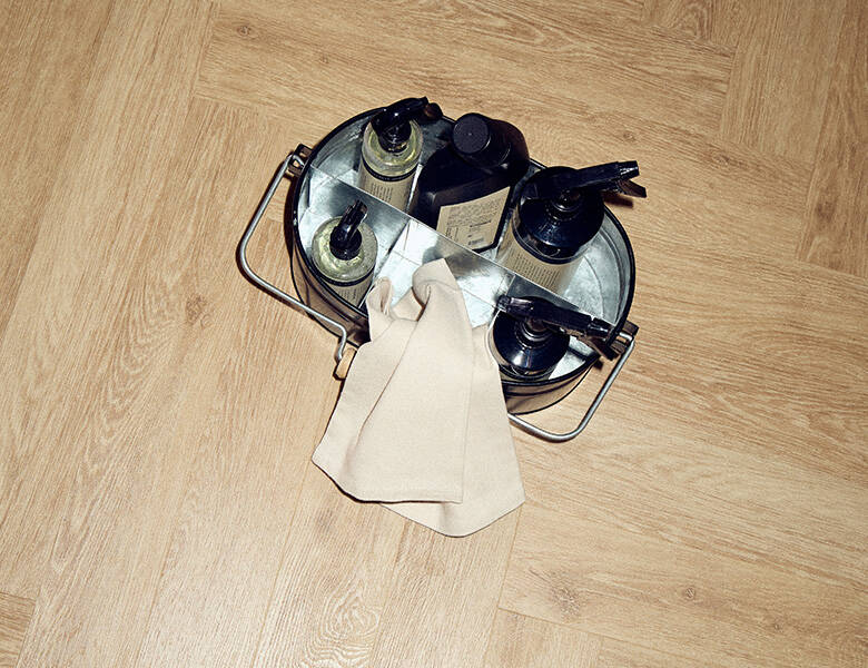 Her ses en spand med forskellige rengøringsting. Den står på et lyst trægulv.