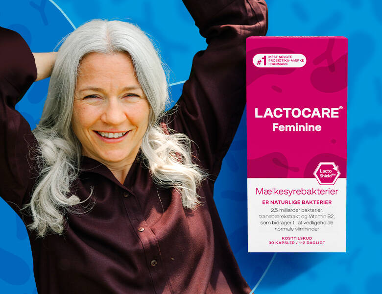 Middelalderlig kvinde med gråt hår iført mørkerød skjorte. Står foran blå baggrund og smiler med armene over hovedet. Der ses Lactocare Feminine product i forgrunden.