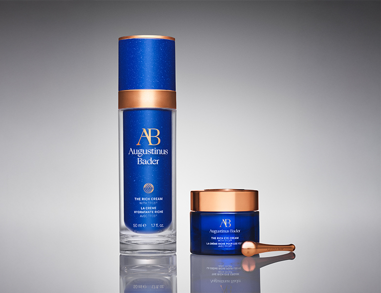 Her ses Augustinus Bader hudplejeprodukter – serum og creme i blå emballage på en mørk baggrund.
