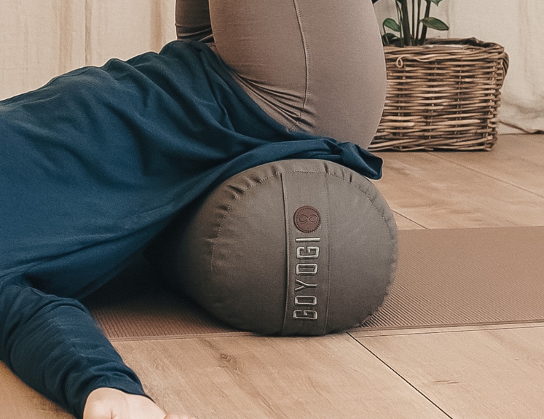 Person ligger på en yogapølle på en måtte i et roligt hjemmemiljø med kurv og plante i baggrunden.