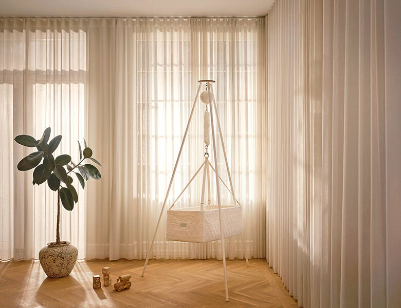 Slyngevugge på stativ i værelse med tynde hvide gardiner trukket for. Trægulv og en stor grøn plante til ventre i en brun-hvid potte. Træklodser på gulvet.