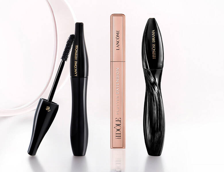 Her ses et billede med tre mascaraprodukter fra Lancôme placeret på en lys, reflekterende overflade mod en lys baggrund. På produkterne står teksten “LANCÔME HYPNÔSE”, “IDÔLE FLUTTER EXTENSION” og “HYPNÔSE DRAMA”. Den ene mascara er vist åben med børsten synlig.