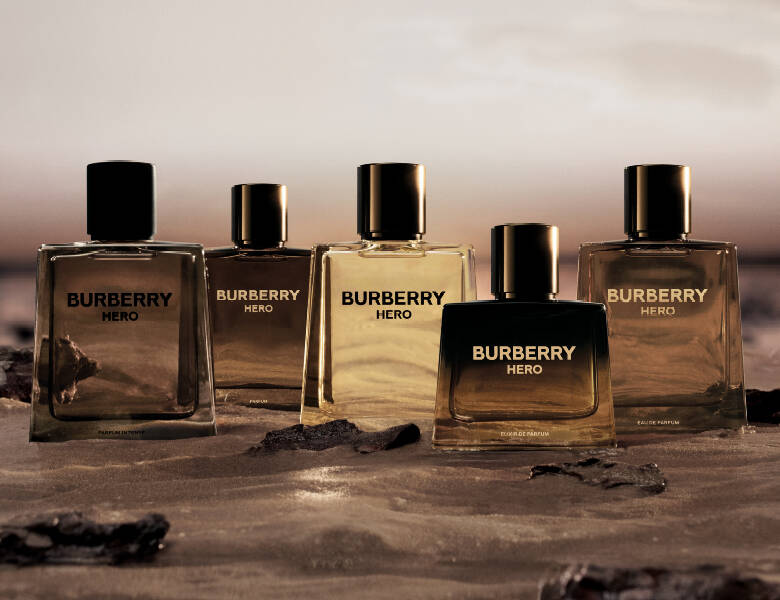 Her ses et billede med flere parfumeflakoner fra Burberry placeret side om side på en sandet overflade med sten. På flakonerne står teksten “BURBERRY HERO”. Flakonerne er i forskellige nuancer fra klar til mørk med sorte låg.