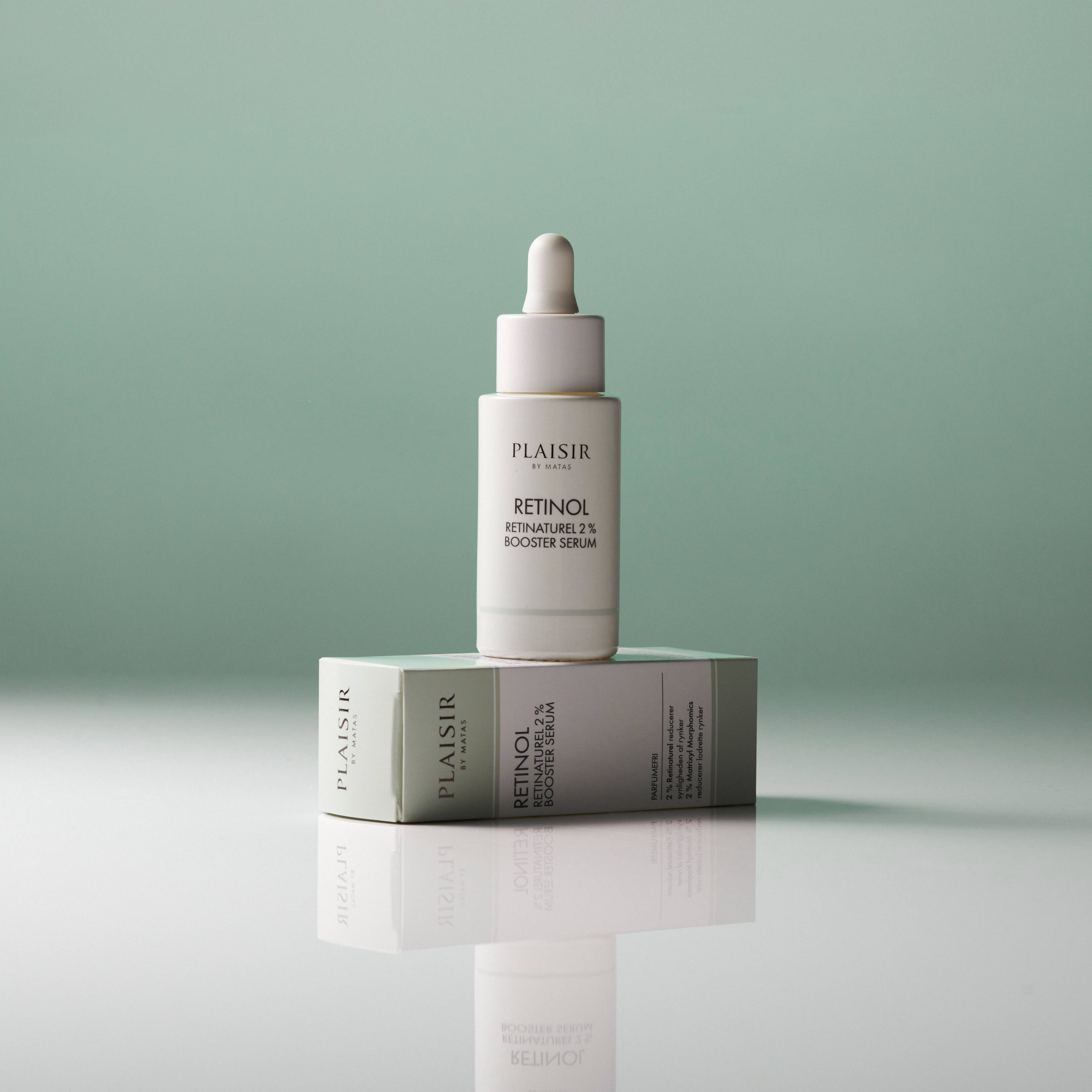 En flaske Plaisir Retinol 2% Booster Serum står oven på sin æske på en blank overflade mod en grøn baggrund. Flasken har pipette og spejler sig i underlaget.