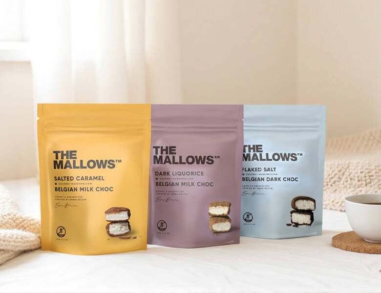 Gul, lilla og lyseblå The Mallows poser i et lyst miljø med en hvid kop med te til højre og et lyst tæppe i baggrunden. De står på et lyst bord