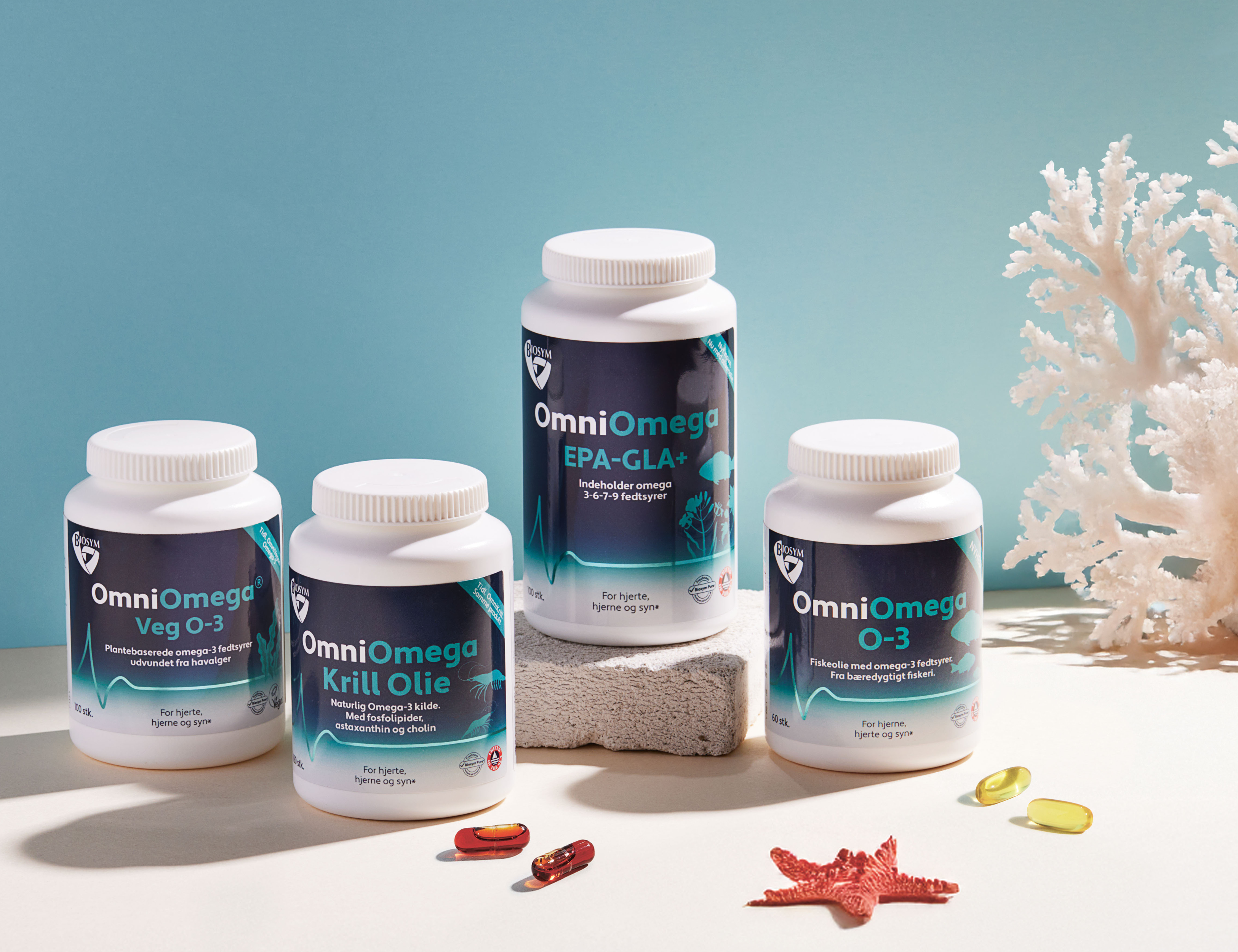 Tre OmniOmega kosttilskud står på en lys overflade foran en blå baggrund sammen med gelkapsler, en rød søstjerne og hvid koral.