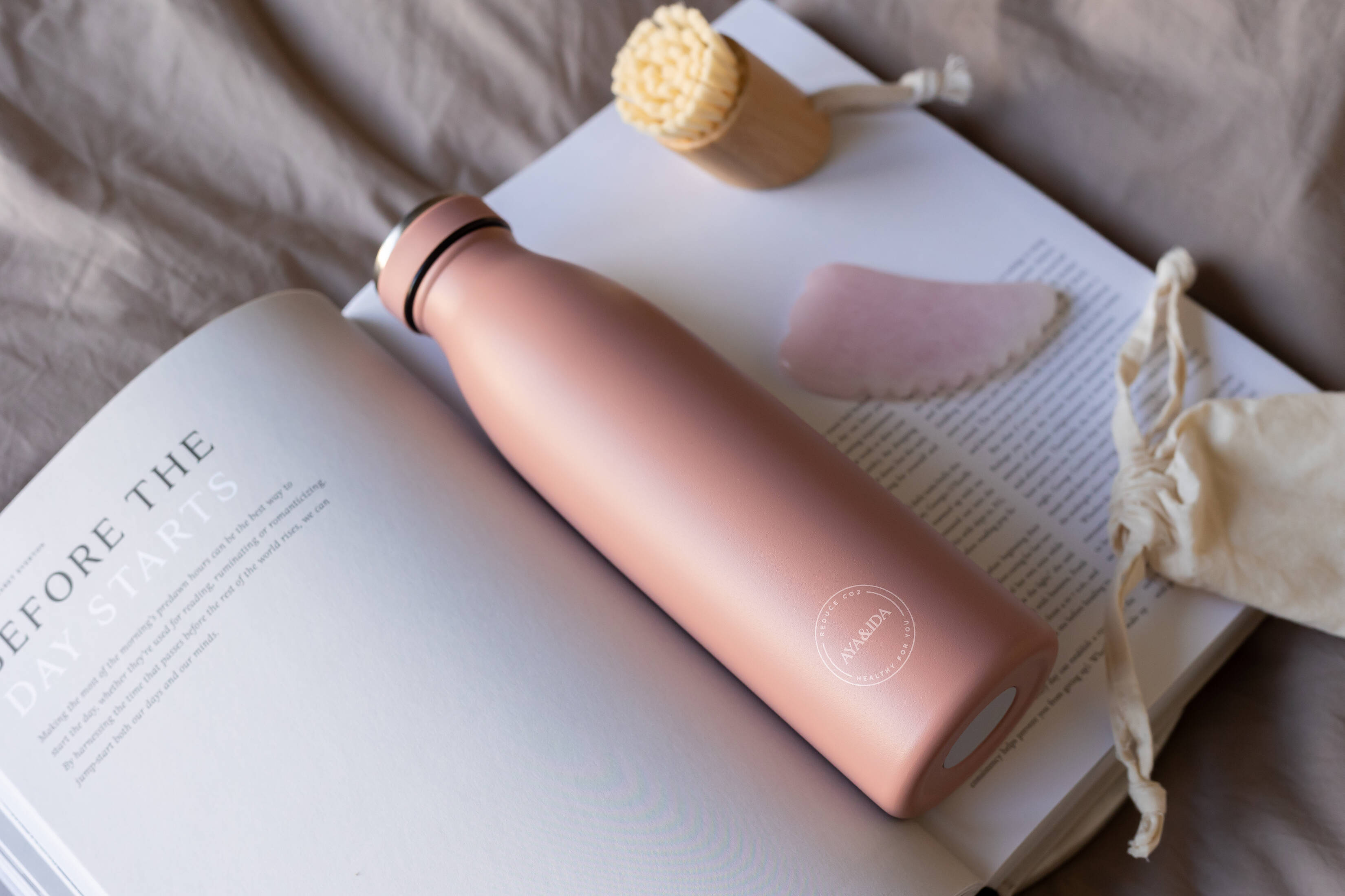 Her ses en lyserød drikkeflaske, som ligger på en åben bog, hvor der også ligger nogle andre produkter. Det hele ligger på en neutral dyne. 