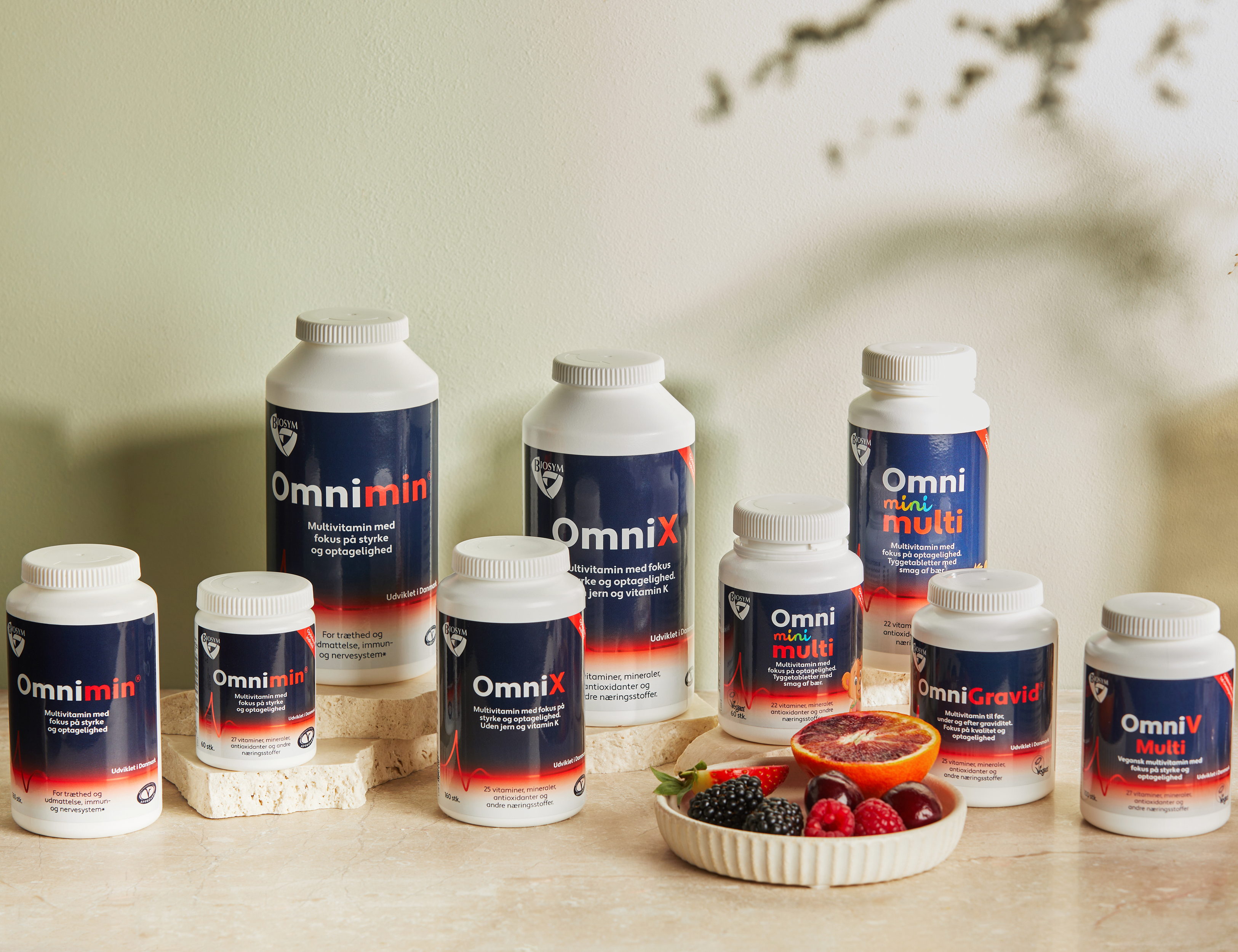 Flere Biosym multivitaminer står samlet på lyse stenplader foran en lys grøn baggrund med en skål fyldt med bær og en halveret blodappelsin.