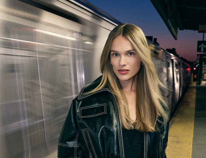 Her ses en kvinde med langt lyst hår. Hun står foran et tog i bevægelse. 