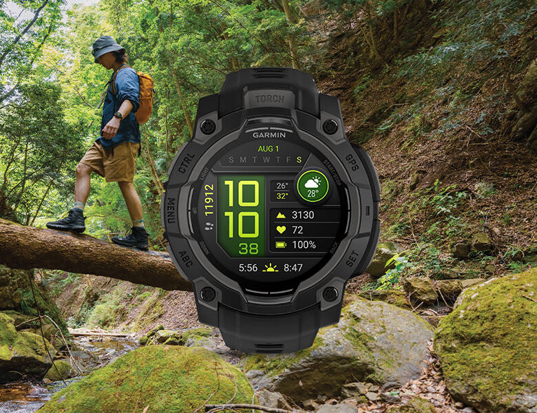 Person vandrer i skoven og balancerer på en træstamme over en bæk, mens et robust Garmin smartwatch med aktivitetsdata vises stort i forgrunden.