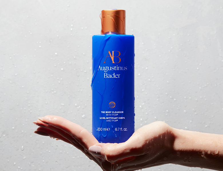 Her ses en hånd, der holder en Augustinus Bader body cleanser i blå emballage med vanddråber på en lys baggrund.