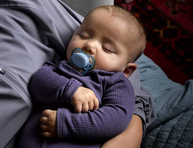 Her ses en baby i en lilla dragt. Babyen sover i armene af en og har en blå sut i munden.